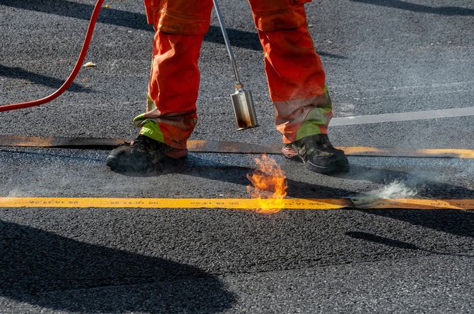 A worker in orange trousers uses a propane torch to melt thermoplastic material onto an asphalt road to create a yellow line.