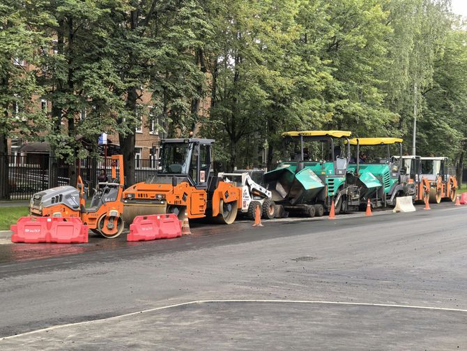 Construction equipment, including rollers and pavers, parked in a row on an asphalt road with red safety barriers.