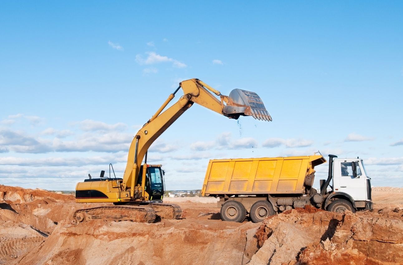 A yellow excavator loading soil into a dump truck at a construction site against a clear blue sky.