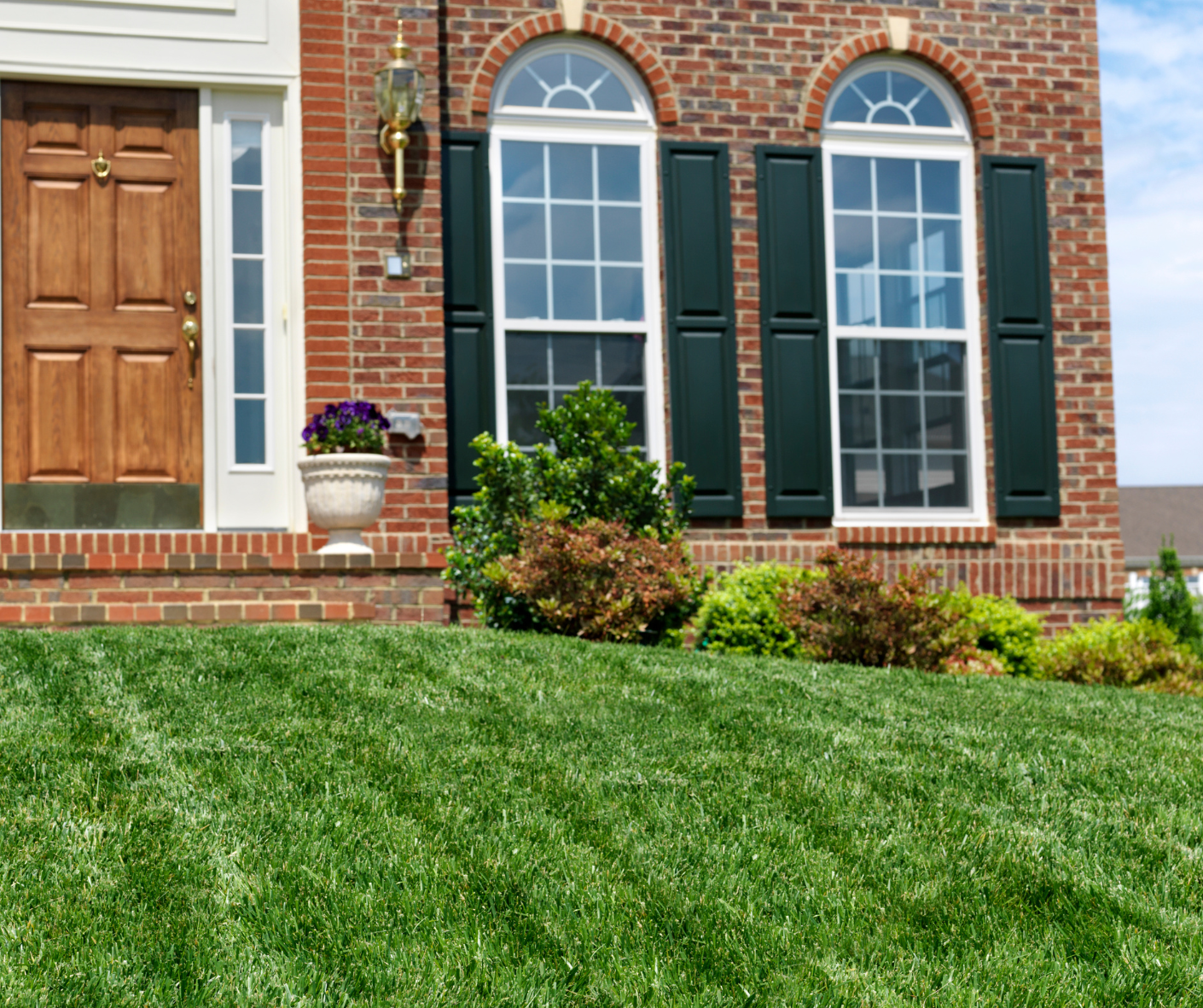 Brick house front entrance with white-trimmed windows, dark shutters, potted flowers, and a green lawn