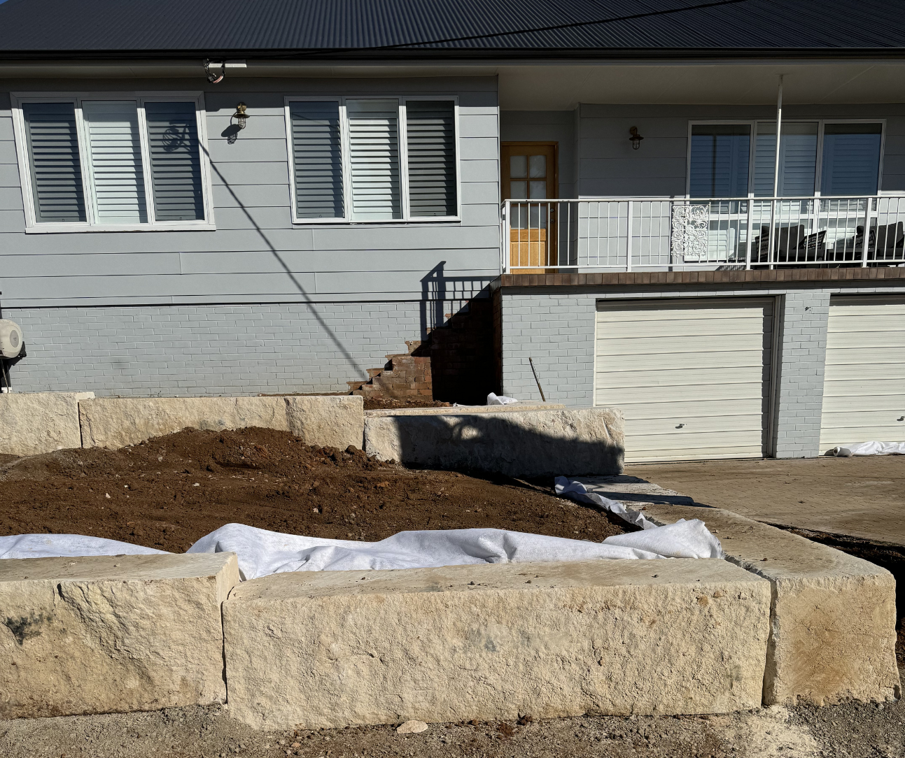 Front yard with exposed soil, landscape fabric, and stone retaining wall in front of a gray house and garage Front yard with exposed soil, landscape fabric, and stone retaining wall in front of a gray house and garage