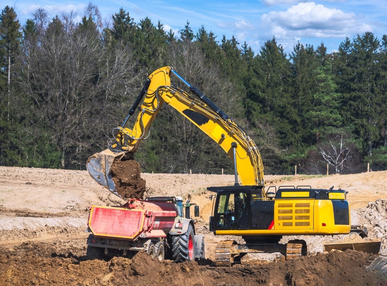 A yellow excavator fills a red dump truck with soil at a construction site in front of a forest.