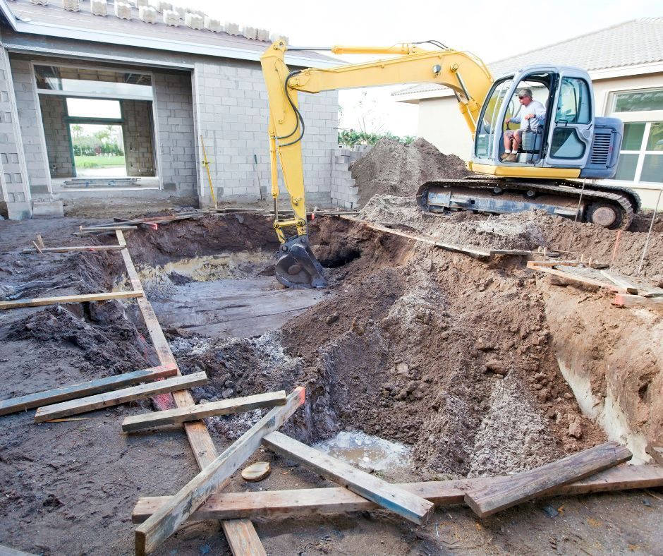 A yellow excavator digging a swimming pool hole in the backyard of a house under construction.