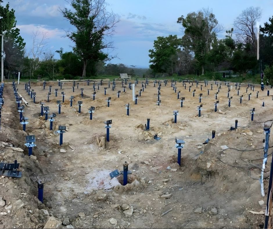 A construction site featuring a grid of numerous blue steel helical piers driven into leveled sandy soil.