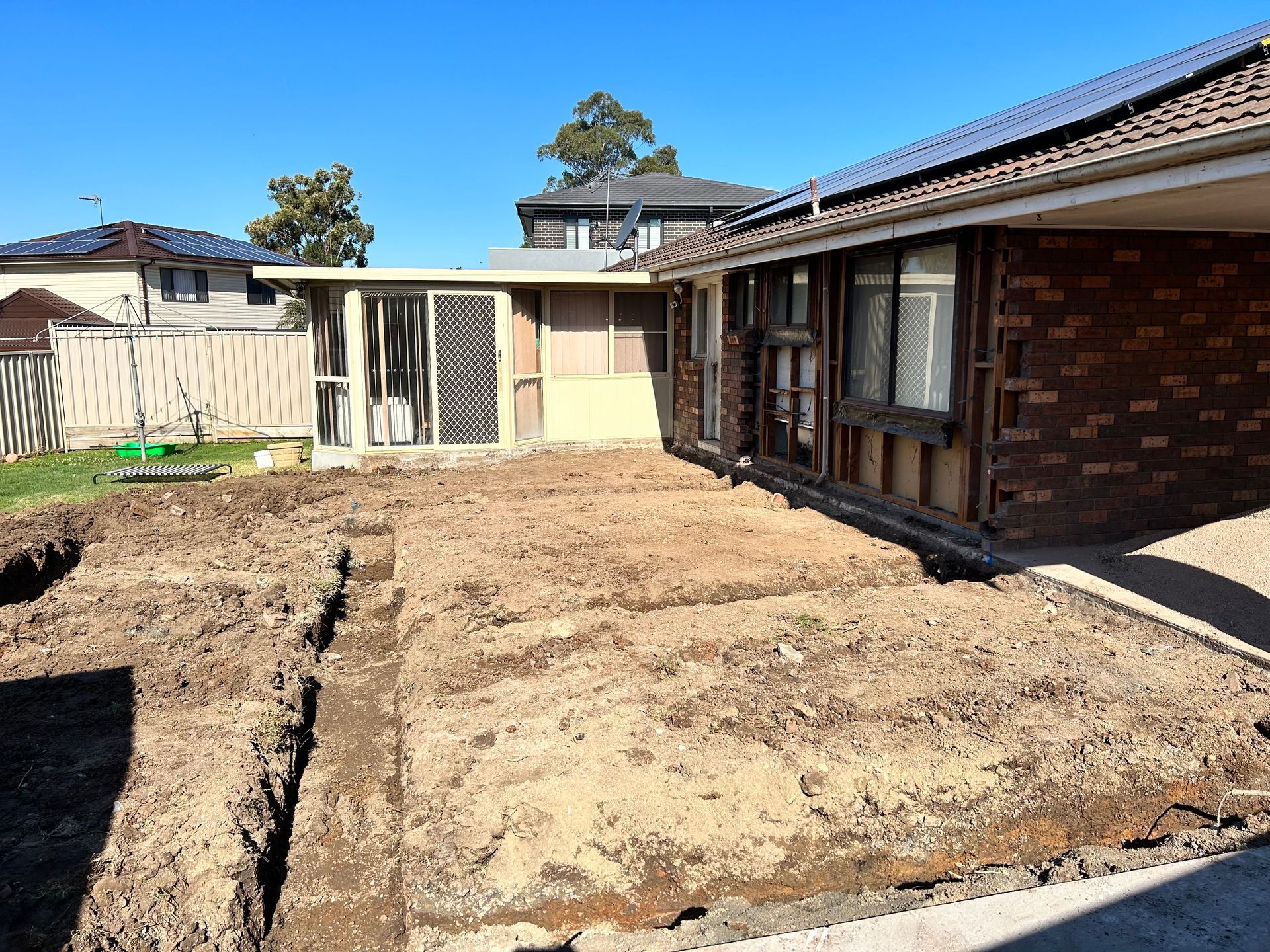 A residential backyard under renovation, featuring a dug-out foundation trench alongside a brick home and fence.