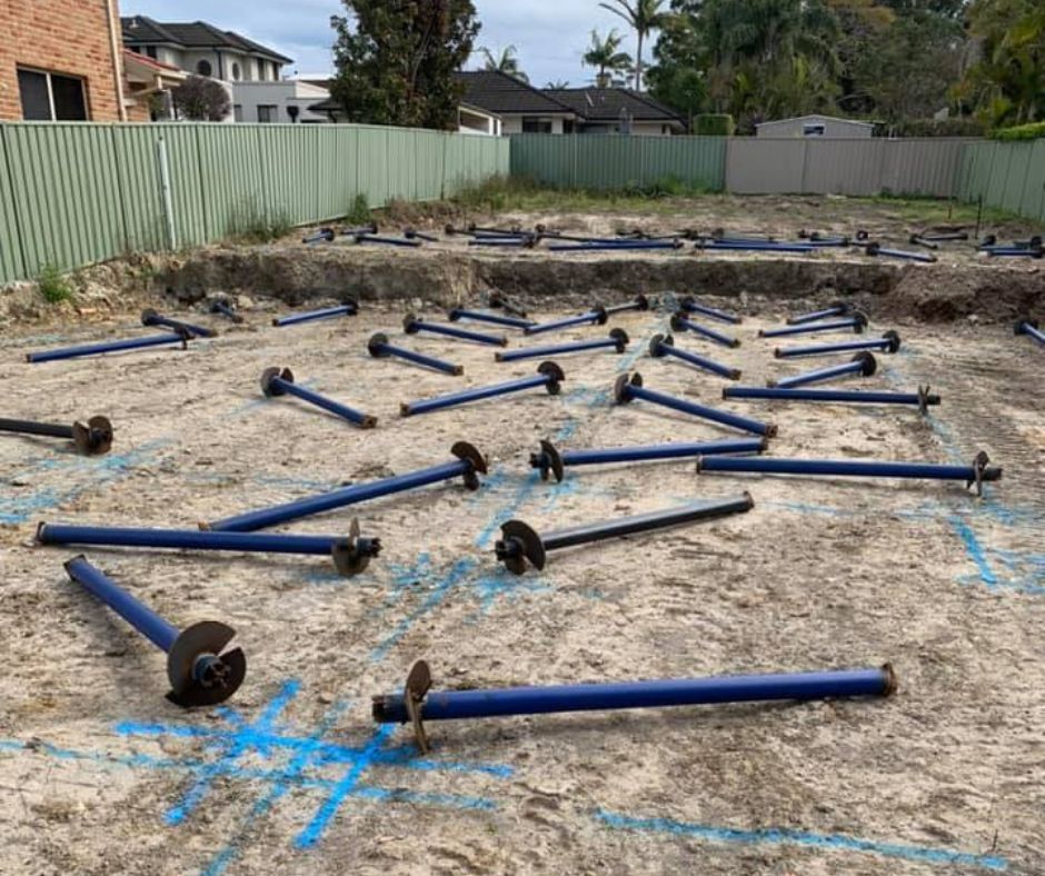 Construction site showing numerous blue screw piles laid out on a dirt ground marked with blue surveyor spray paint.