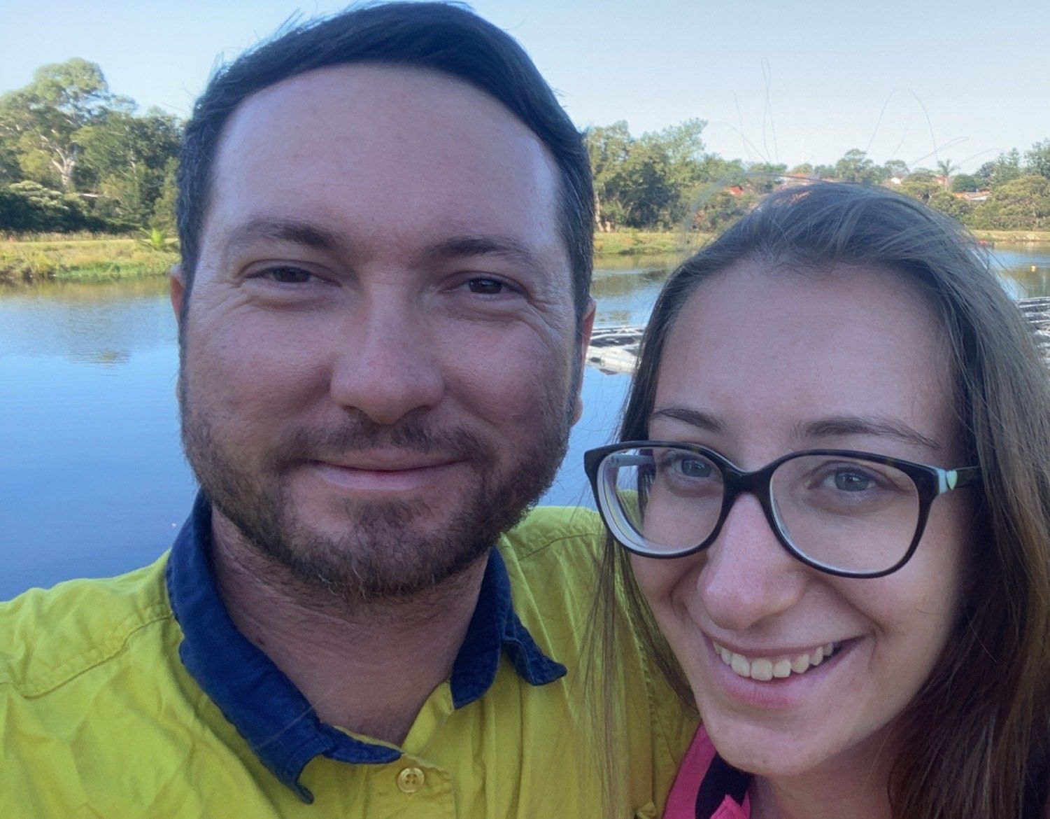 A smiling couple poses for a selfie outdoors with a body of water and trees in the background.