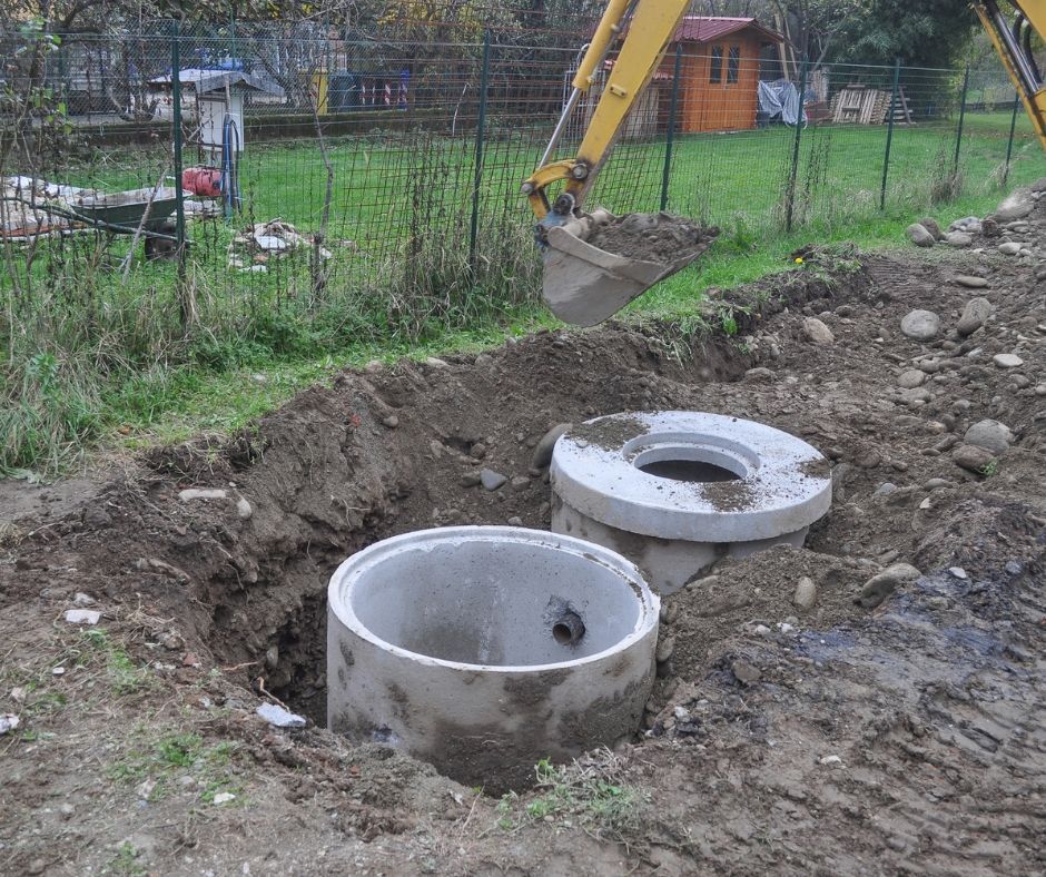 An excavator bucket hovers above two circular concrete sewer manhole sections installed in a grassy residential yard.