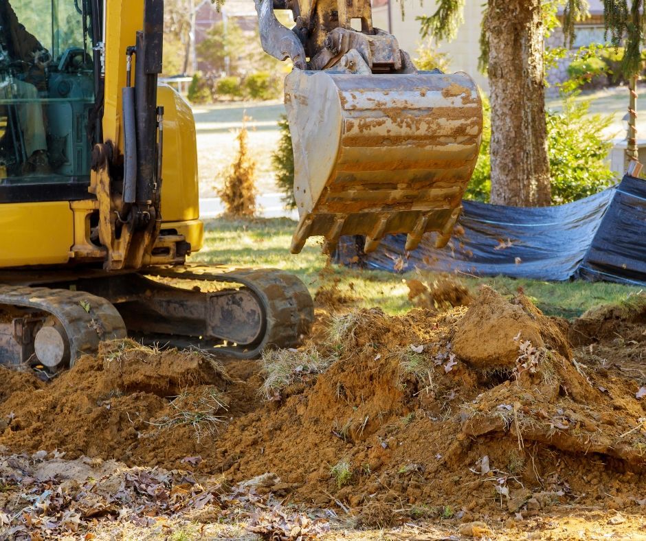 A yellow excavator with its bucket extended over a pile of loose, brown dirt in a grassy residential area.