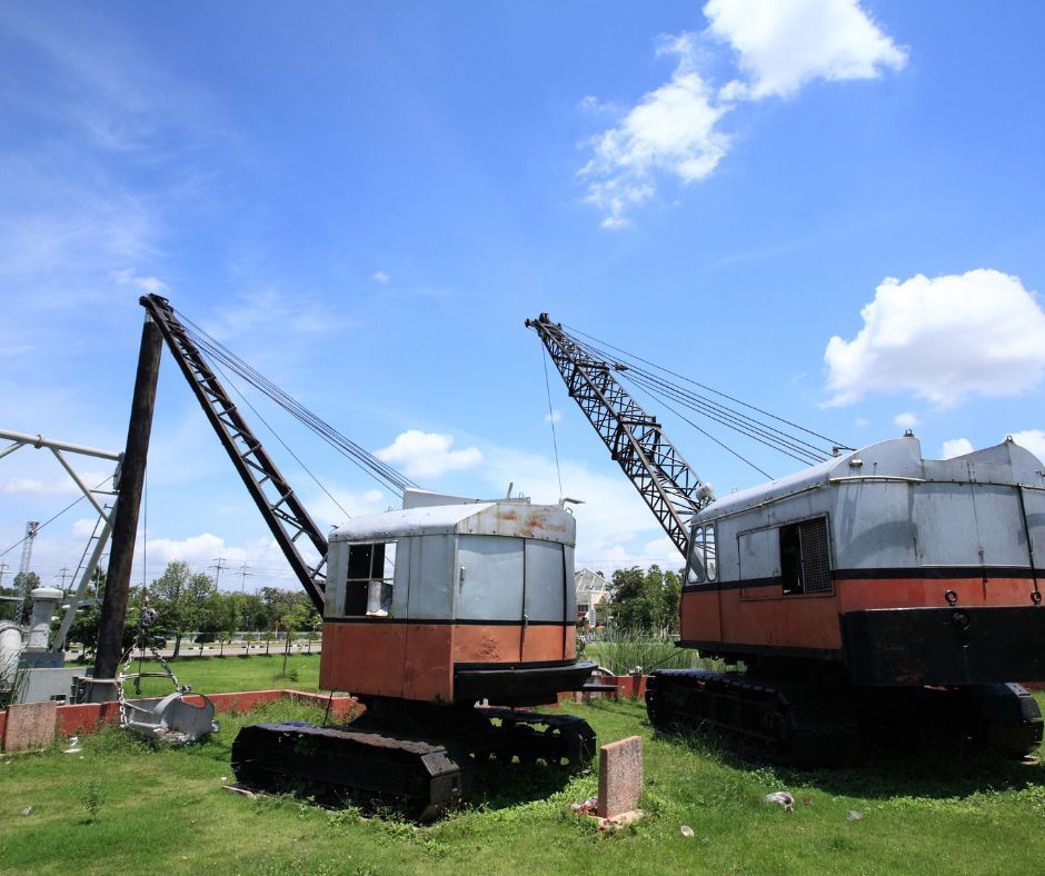 Two vintage, orange and silver crawler cranes parked in a grassy field under a bright, sunny blue sky.