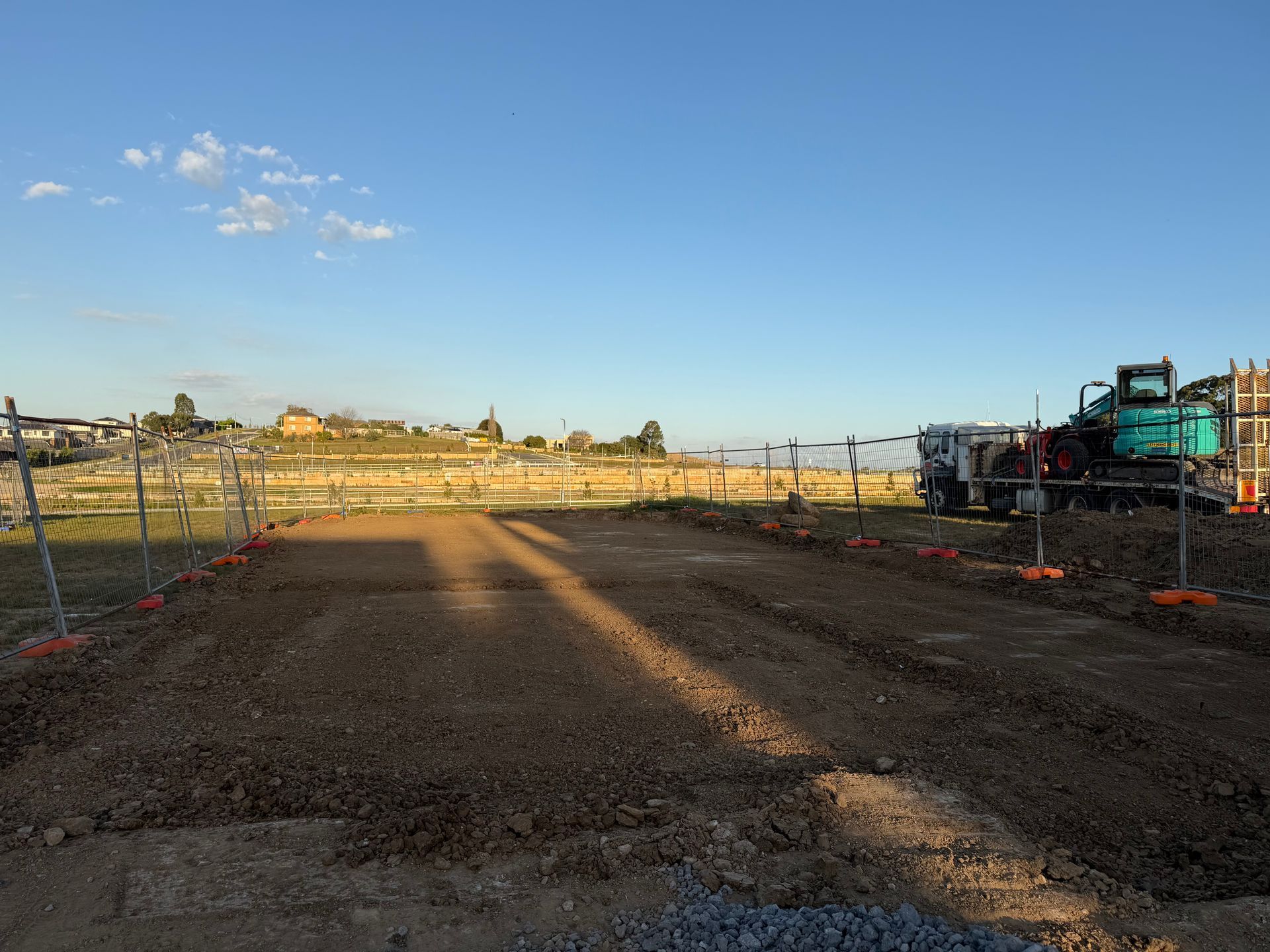 Aerial view of a teal excavator digging a trench in a fenced dirt construction site next to a road.
