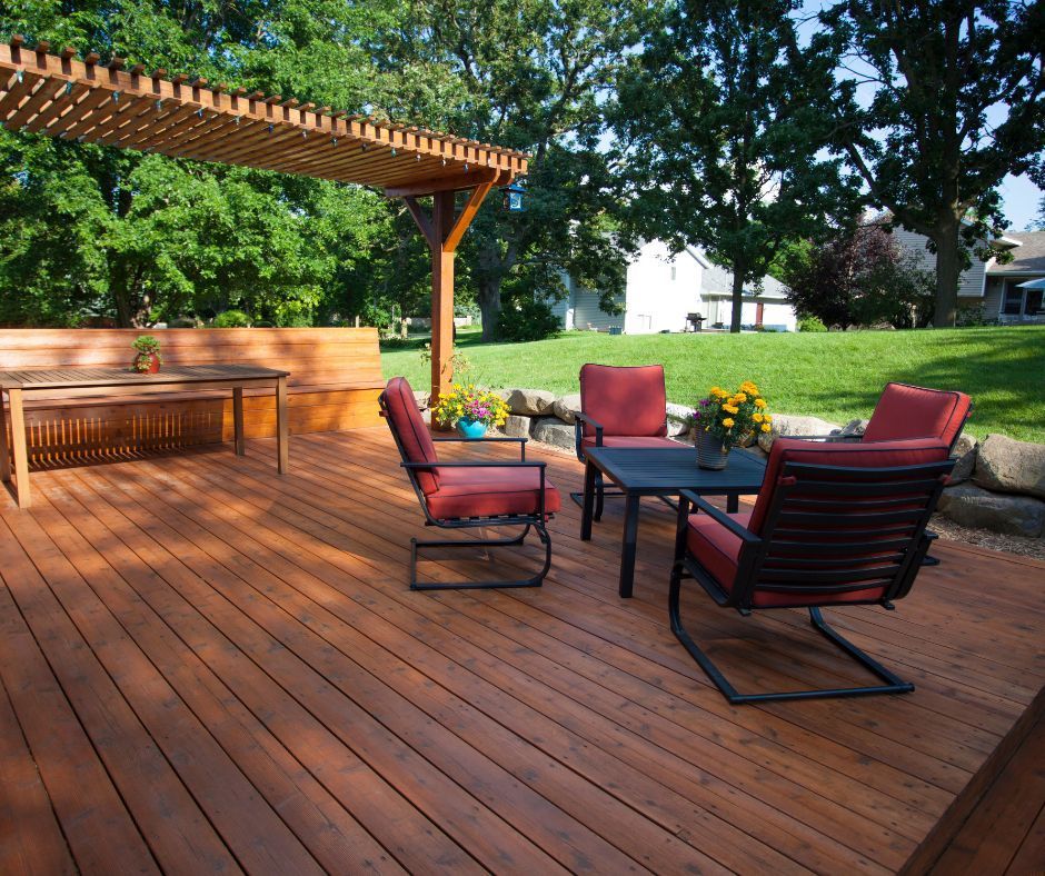 A sunny wooden deck with a pergola, a bench, a small table, and four cushioned chairs in a backyard garden setting.