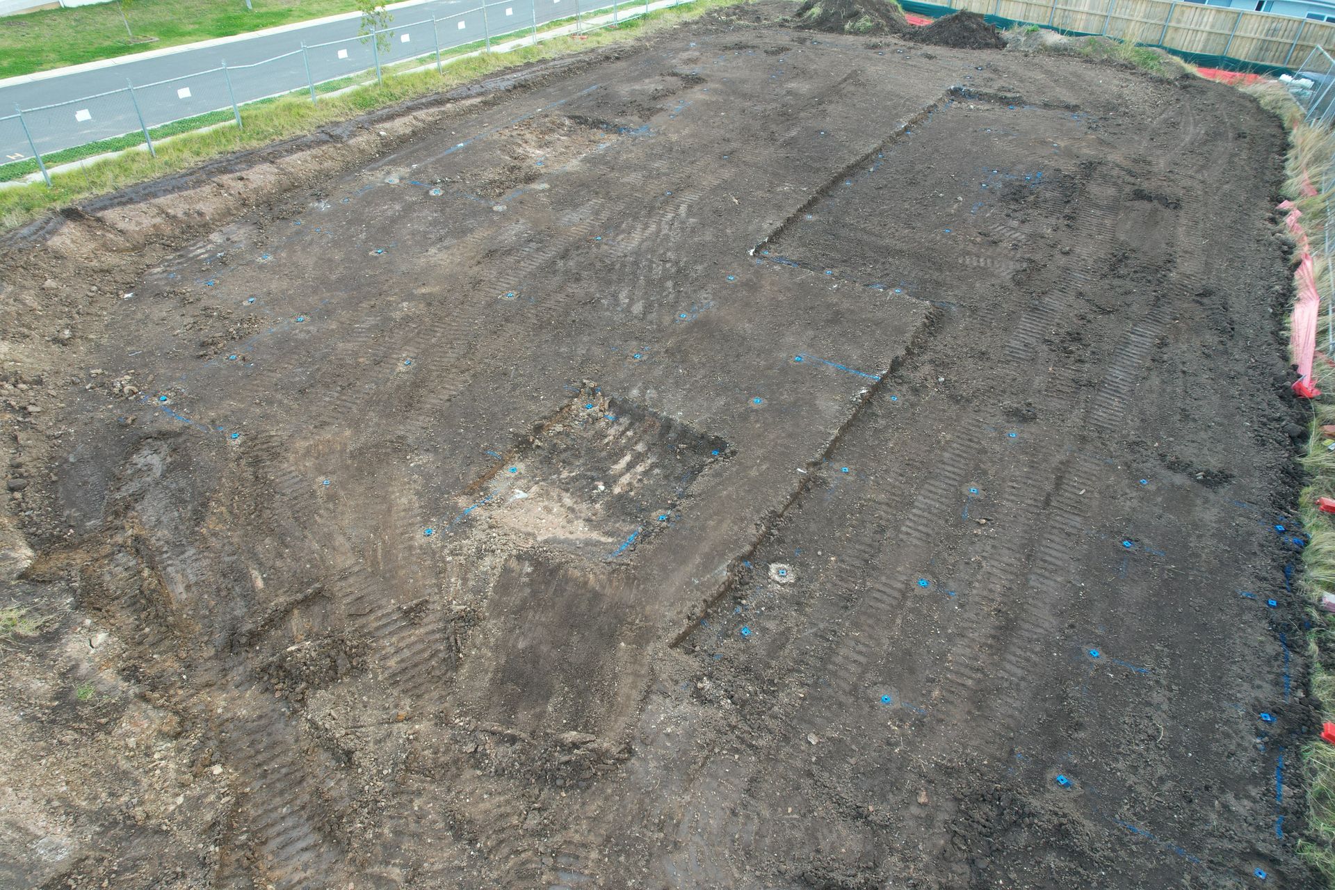 An aerial view of a rectangular construction site with freshly excavated, dark brown soil next to a paved path.