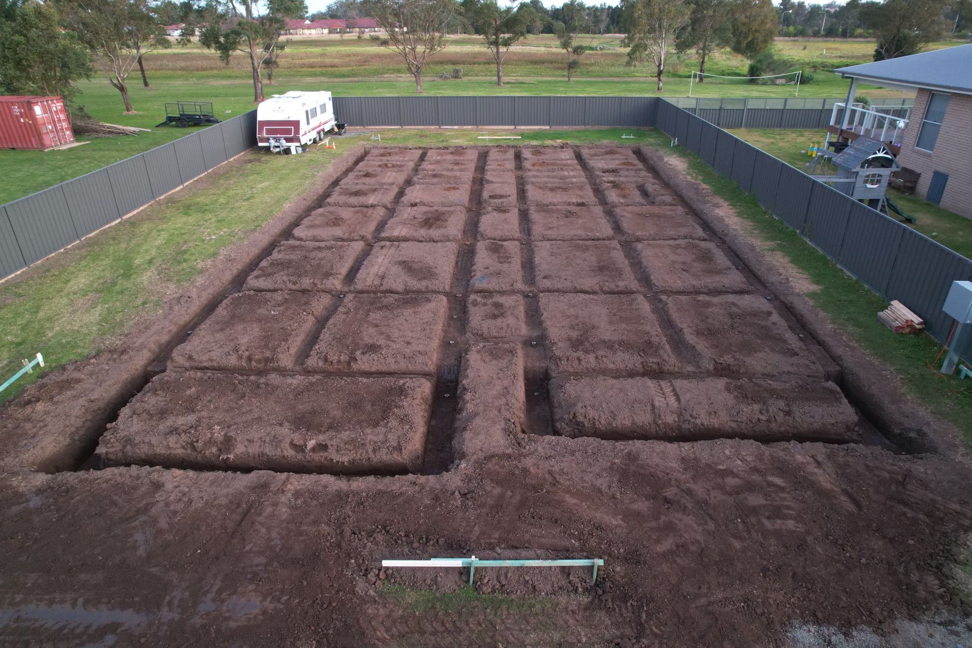 An aerial view of a backyard showing a large, rectangular grid of trenches excavated in the soil for a building foundation.