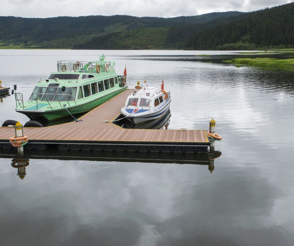 A green passenger ferry and a smaller white boat docked at a wooden pier on a calm lake surrounded by forested hills.