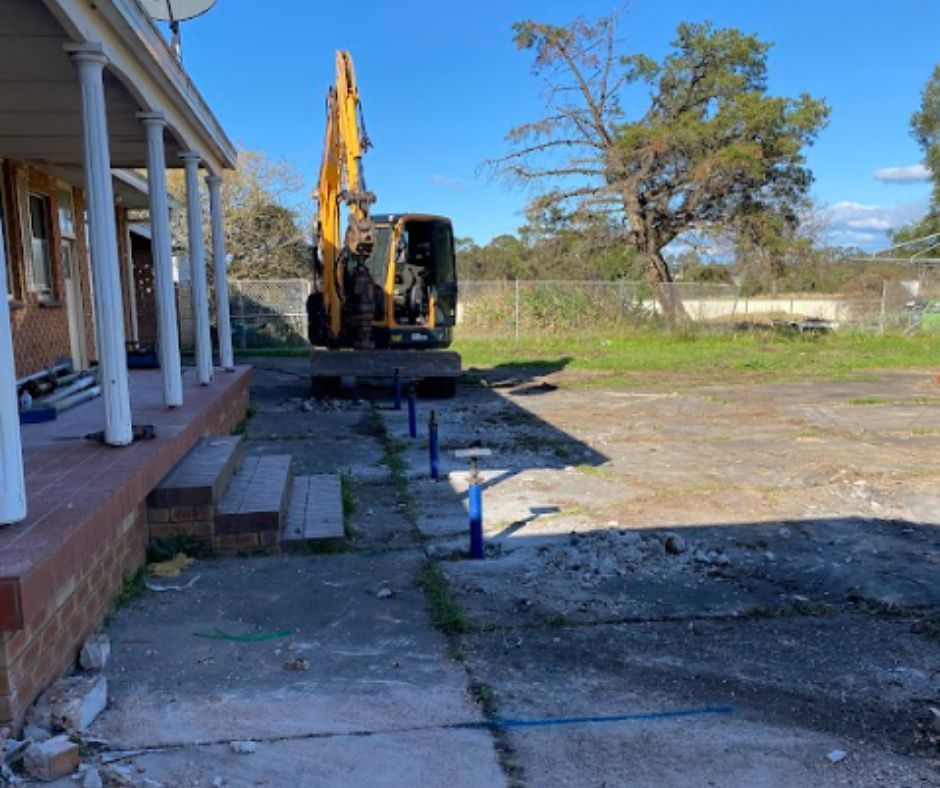 Yellow excavator parked near a house porch with a line of blue construction markers on the ground.