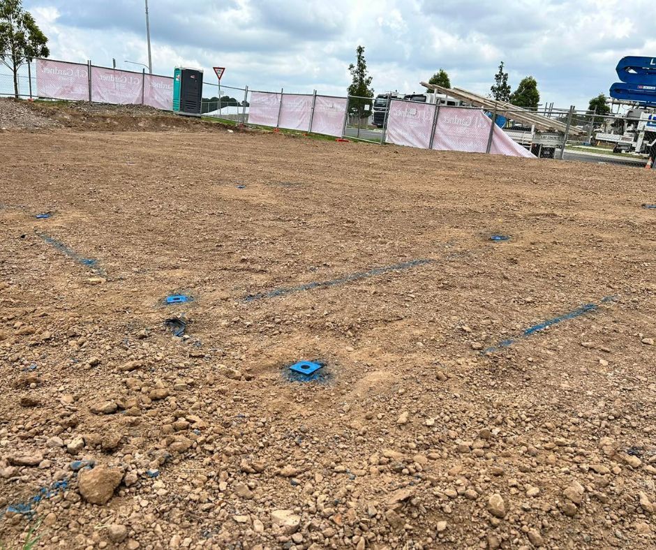 A construction site shows blue metal survey markers and spray-painted layout lines on a dirt-covered ground.
