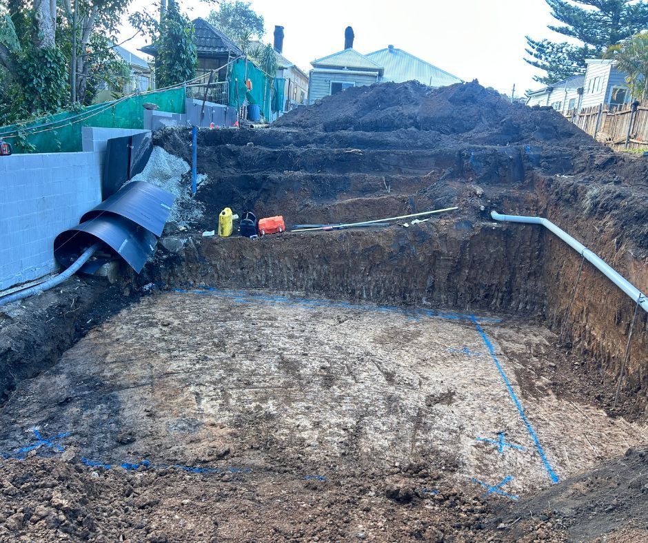 A deep, rectangular excavation in the ground for a pool, with exposed dirt walls, blue spray-painted outlines, and pipes.