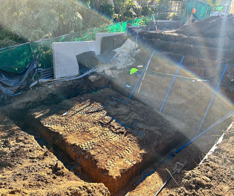 An excavated square trench in a dirt construction site with blue marker poles and white retaining wall blocks.