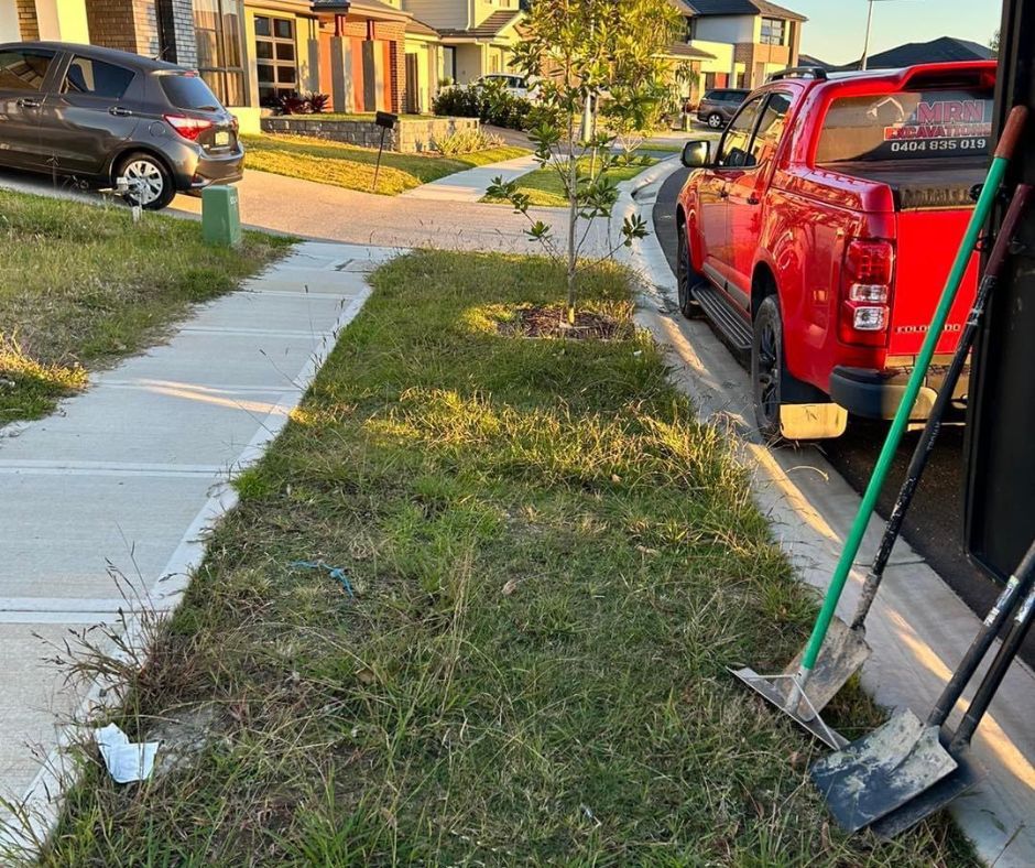 A grassy residential verge with a sidewalk on the left, a red pickup truck on the right, and two shovels leaning nearby.