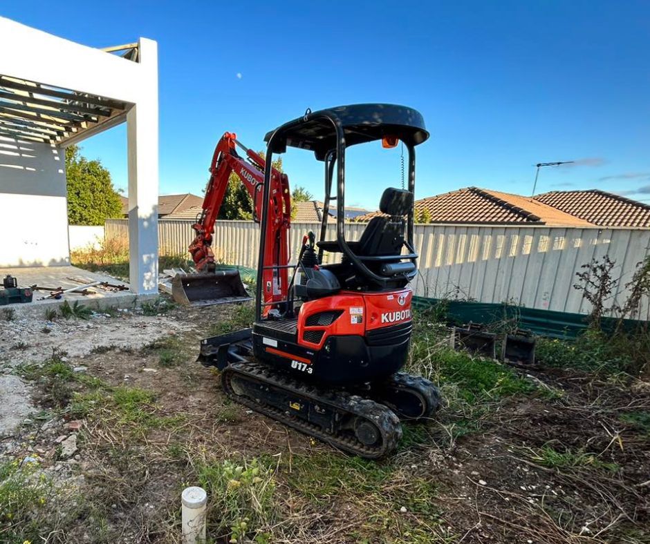 A red and black Kubota compact excavator parked in a backyard construction site under a clear blue sky.