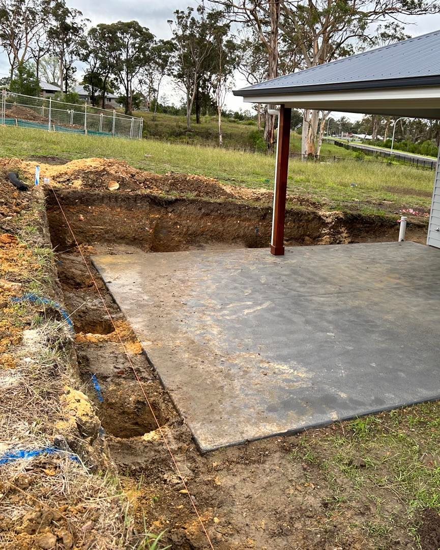 A concrete slab next to a newly excavated soil trench near a building with a partial metal roof.
