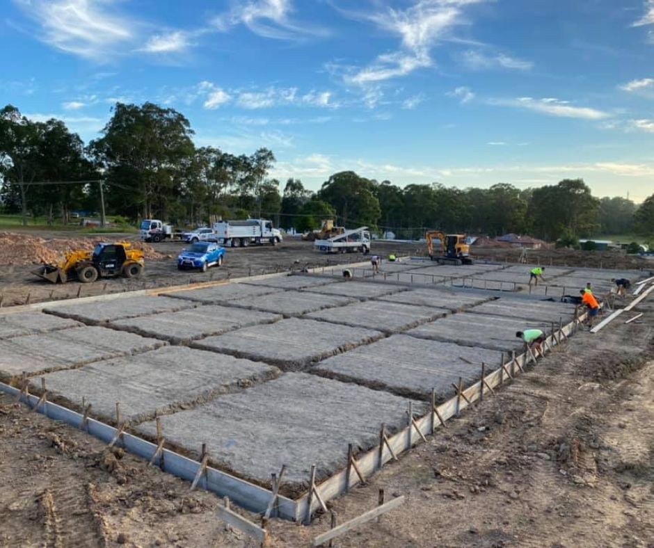 Workers prepare a concrete building foundation with metal grid reinforcements at a construction site under a blue sky.