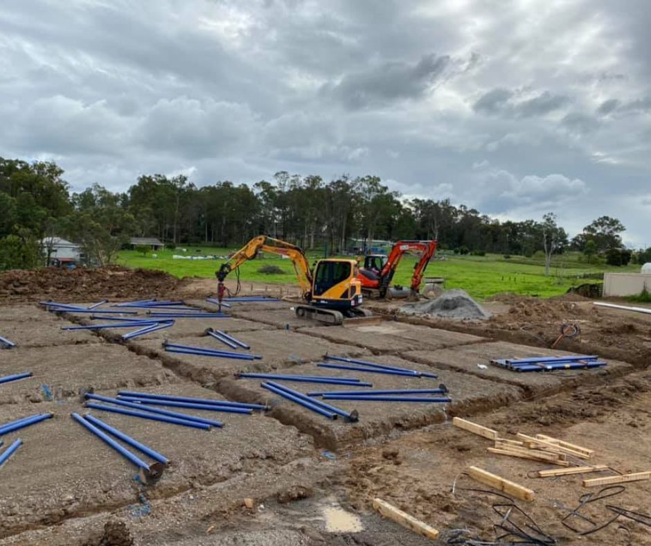 An excavator works on a construction site with blue metal bracing beams arranged in a grid across the dirt ground.