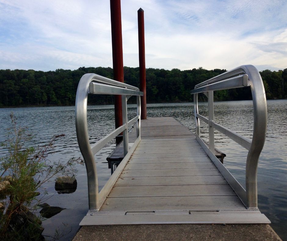A metal-railed ramp leads onto a wooden dock over a calm lake, with two tall poles and a treeline in the distance.