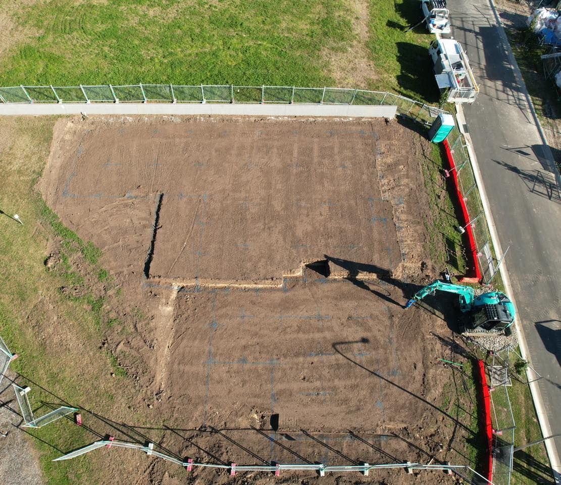 Aerial view of a teal excavator digging a trench in a fenced dirt construction site next to a road.