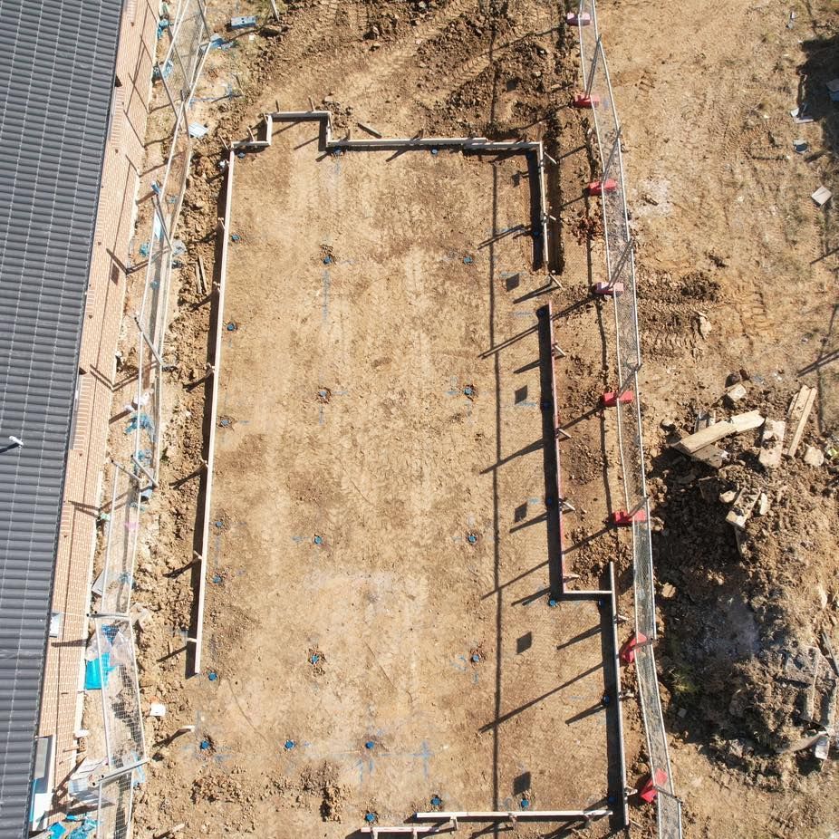 An aerial view of a rectangular building foundation under construction, featuring concrete footings set in bare dirt.