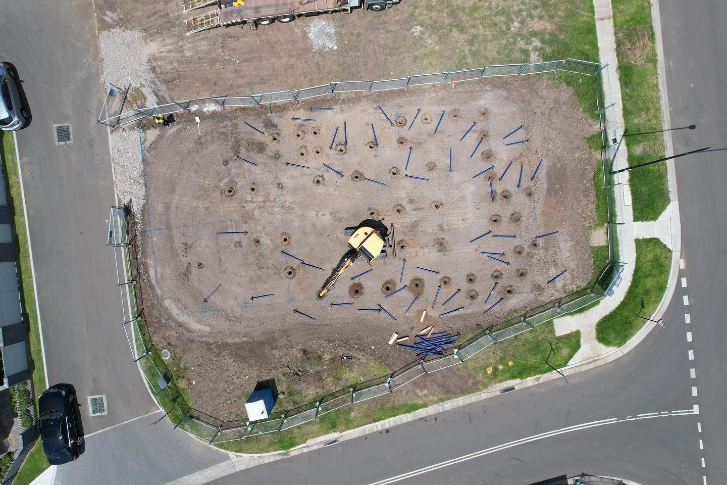 Aerial view of a construction site with a yellow excavator surrounded by numerous small holes in a dirt lot.