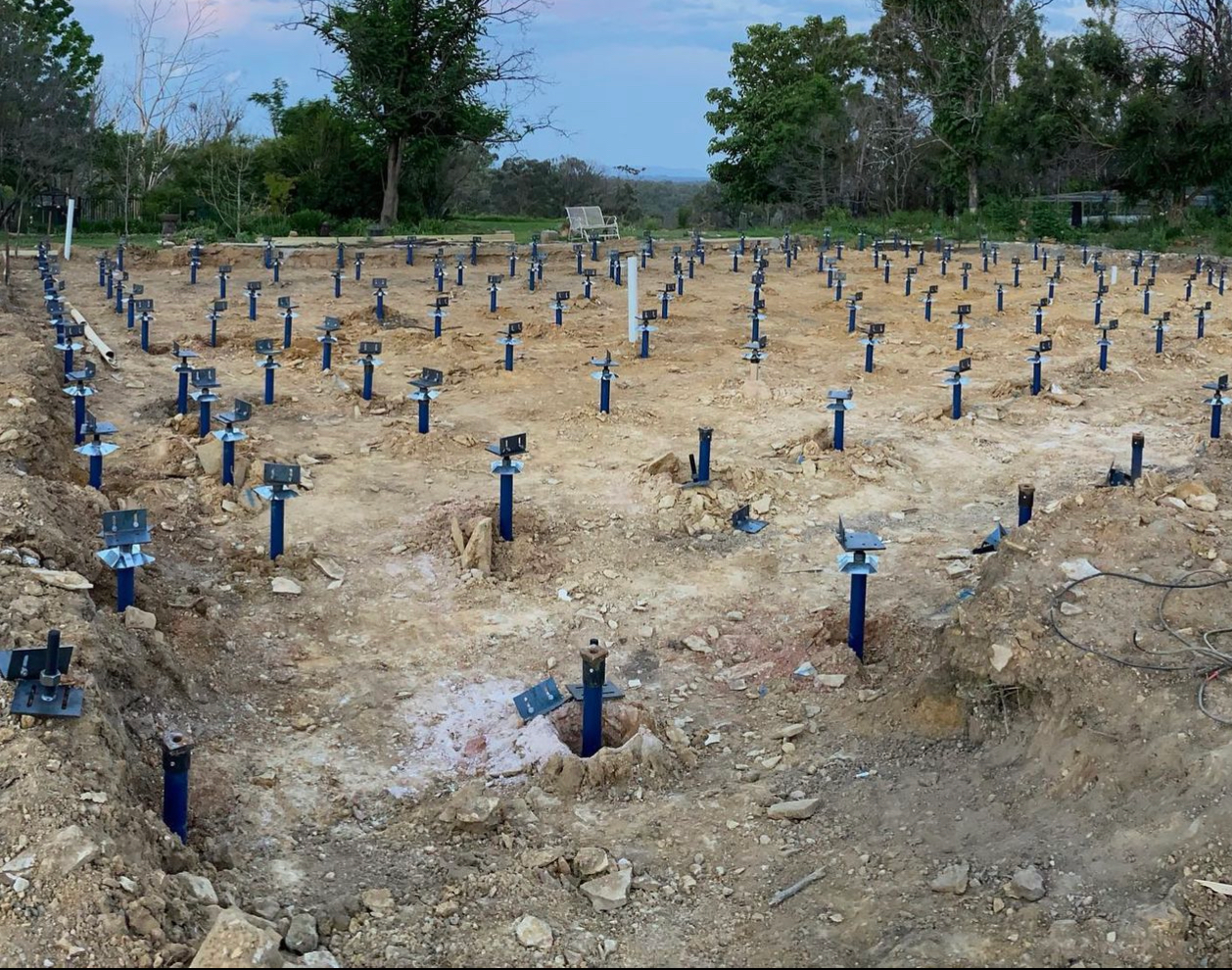 Rows of blue metal stakes in a cleared dirt field, with trees in the background.