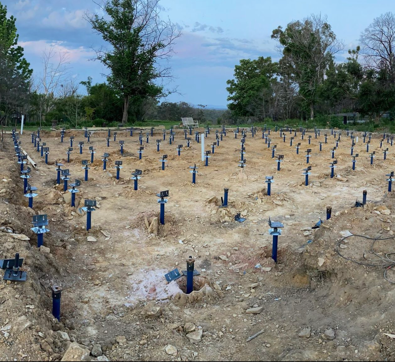 A construction site featuring a grid of numerous blue steel helical pile foundations installed in sandy, excavated earth.
