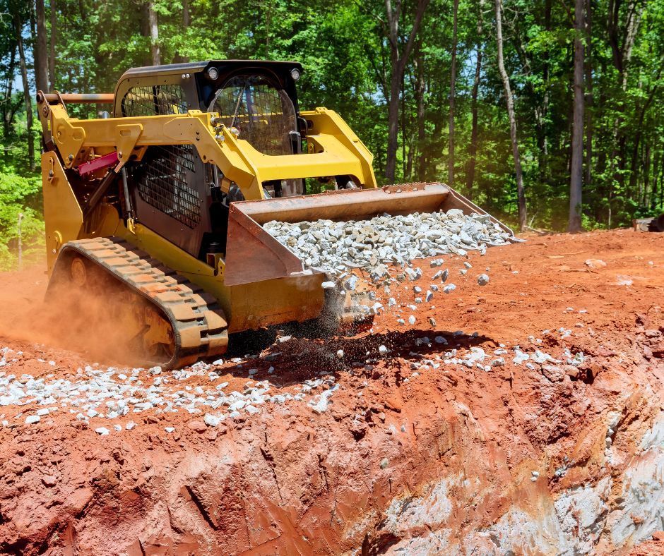 A yellow compact track loader spreading gravel over red clay soil at a construction site near trees.