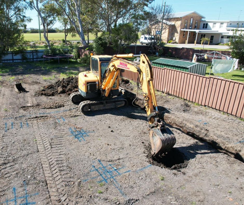 A yellow excavator digs a trench in a dirt yard marked with blue layout lines near a fence and house.