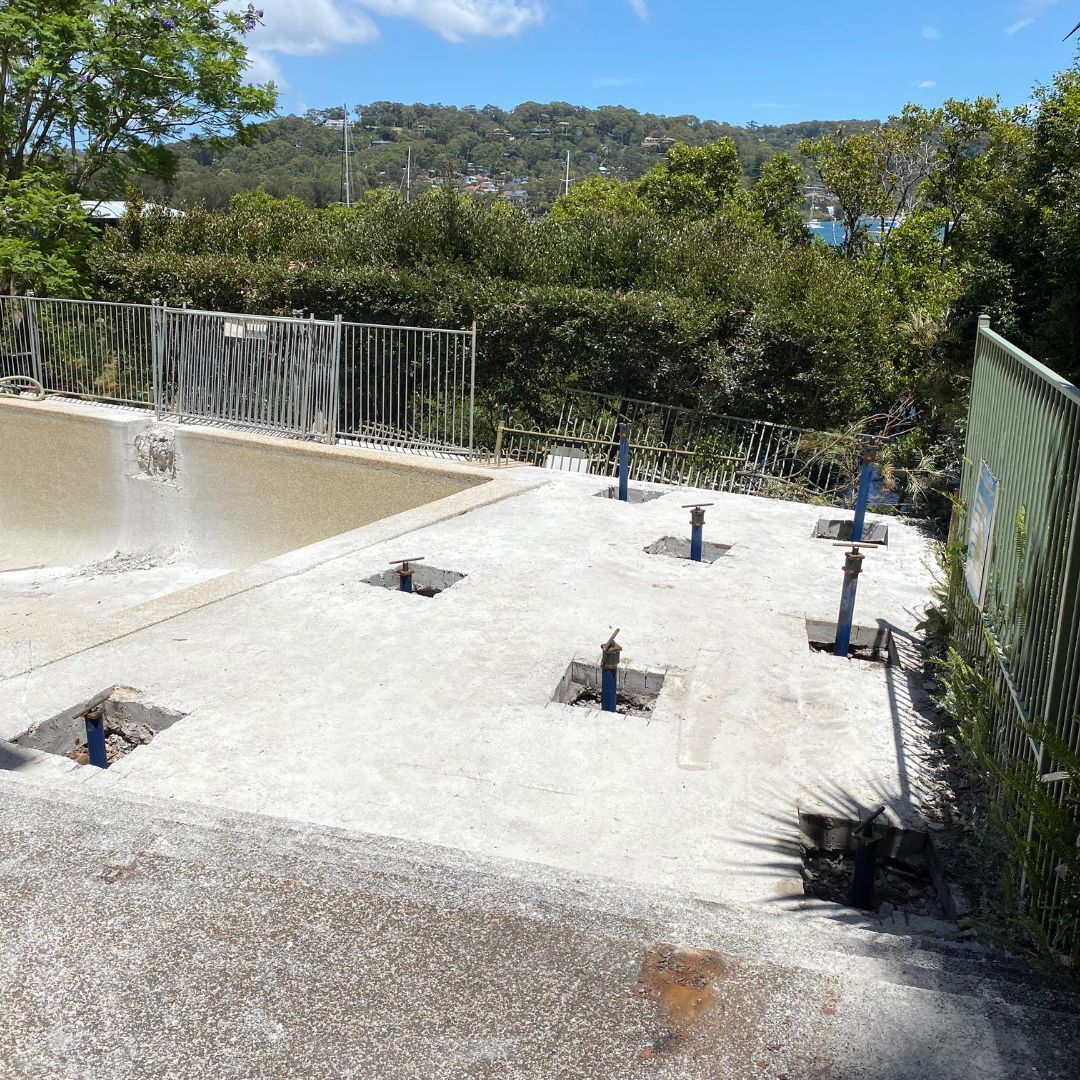 A concrete pool deck with several metal anchor posts sticking up, next to an empty swimming pool.