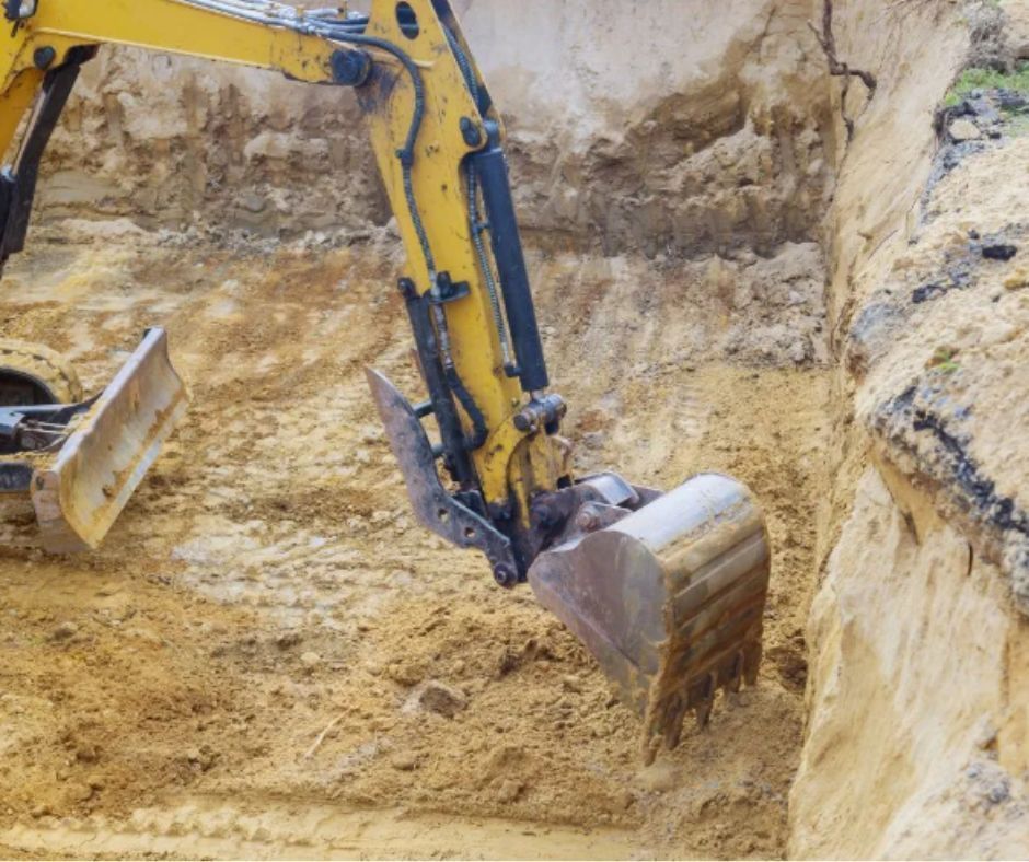 A yellow excavator arm and bucket digging into sandy earth at a construction site.