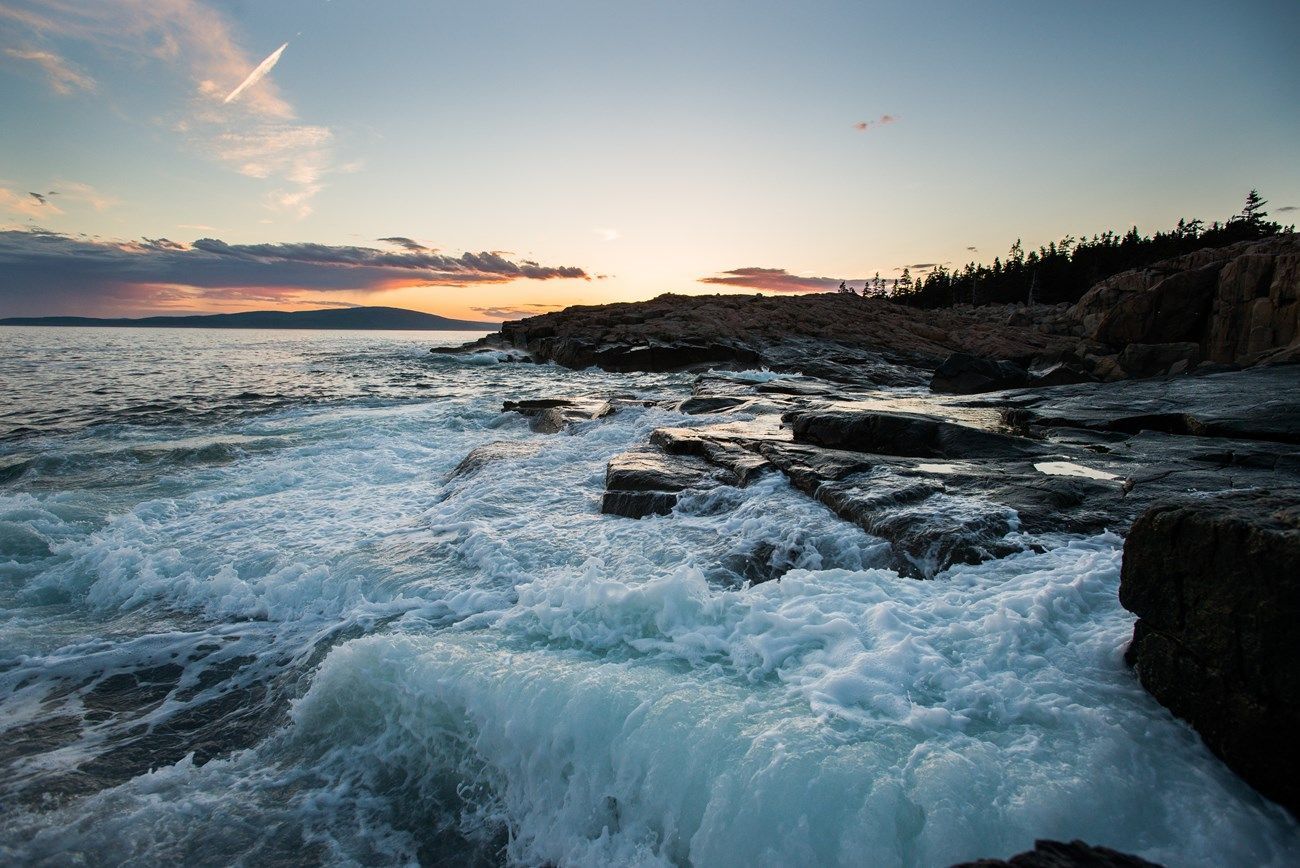 Waves crashing against a rocky coastline at sunset. The sky has streaks of pink and orange.