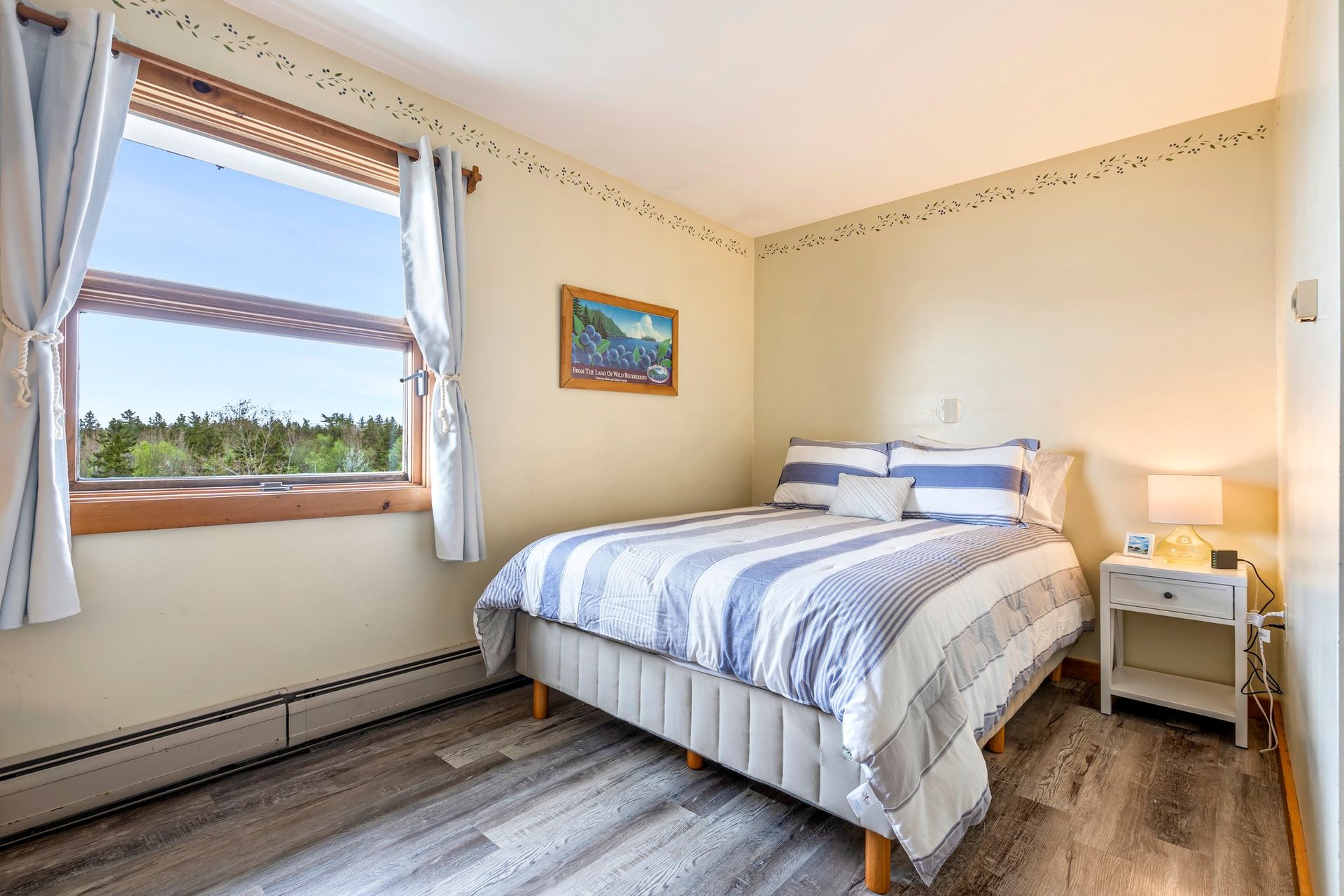 Cozy bedroom with a bed, nightstand, and large window with a view of trees. Blue and white striped bedding.