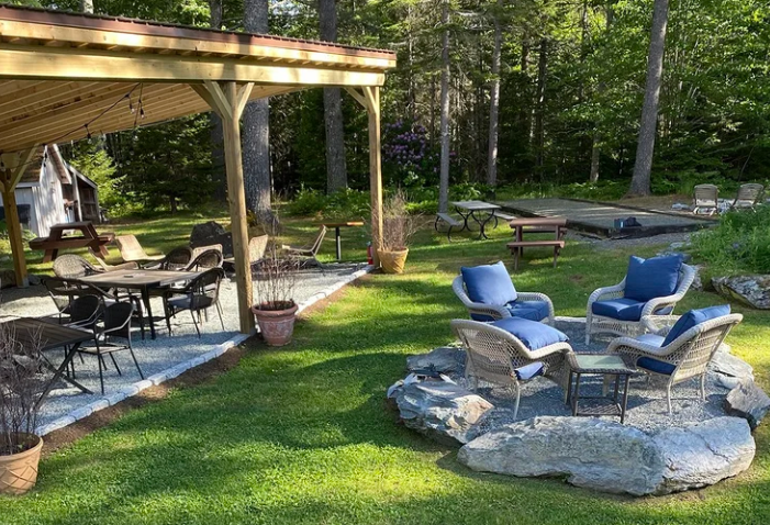 Ironbound backyard patio with blue chairs around a stone fire pit, picnic tables, and a covered deck in a wooded yard
