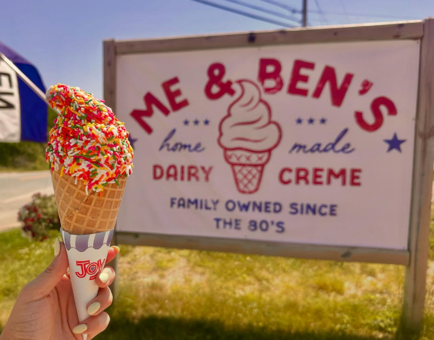 Hand holding sprinkled ice cream cone before a Me & Ben’s Dairy Creme sign