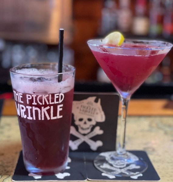 Two pink cocktails on a bar, one in a skull-marked glass with a black straw and lemon garnish.