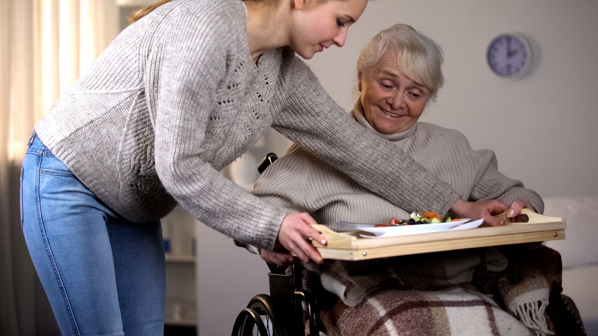 A woman is helping an elderly woman in a wheelchair with a tray of food.