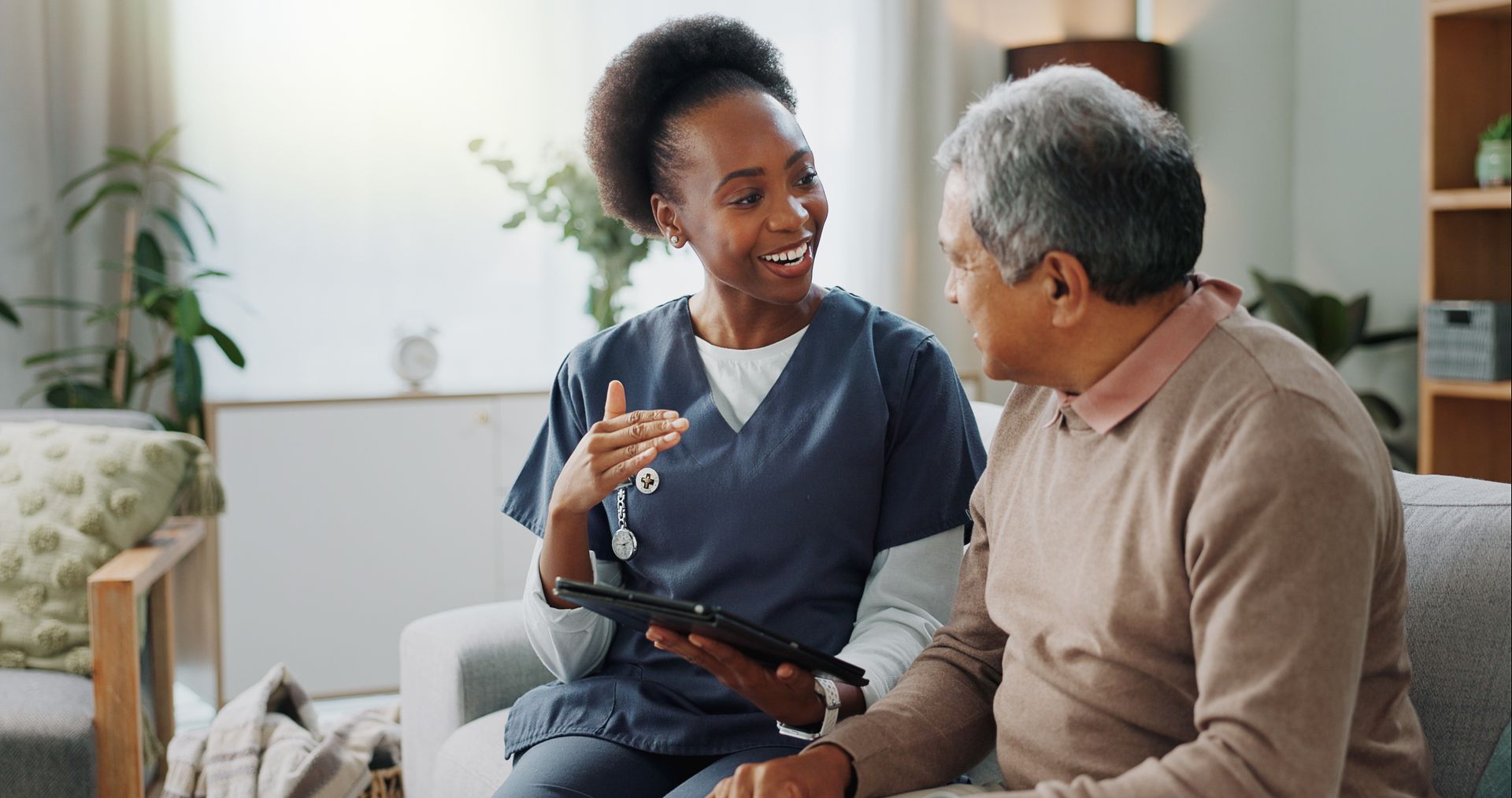A nurse is talking to an elderly man on a couch while holding a tablet.