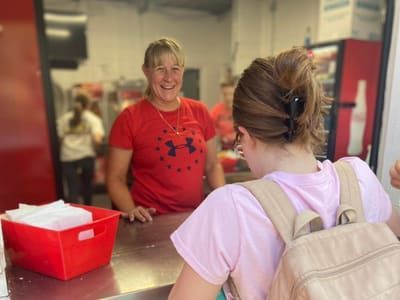 Woman in red shirt smiles, serving food to a person with a pink backpack in a food service area.