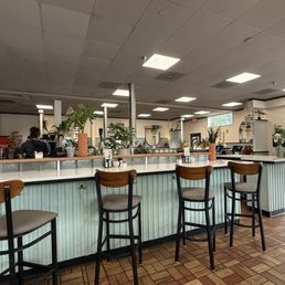 Empty café counter with bar stools, potted plants, and a tiled floor under bright ceiling lights