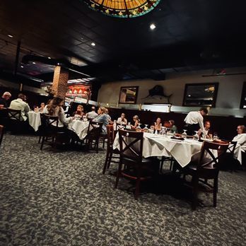 Dimly lit restaurant dining room with diners seated at white-clothed tables and a patterned carpet.