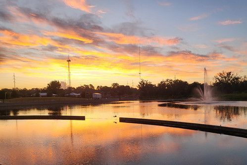 Sunset over a calm pond with fountain, silhouetted trees, and orange-pink sky reflecting on the water