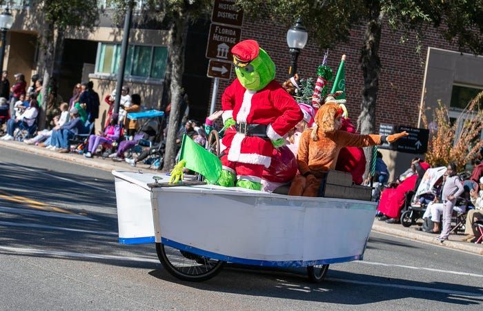 Person in a red Santa outfit and green Grinch mask rides in a white parade float cart on a city street.